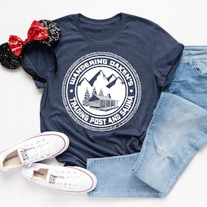 May include: A navy blue t-shirt with a white circle graphic that says "Wandering Oaken's Trading Post and Sauna" with a cabin and mountains inside the circle. The t-shirt is laid out on a white surface with a pair of white sneakers and blue jeans.