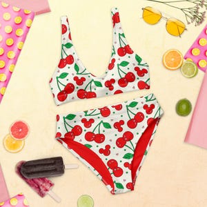 May include: A two-piece bikini set with a white background and a pattern of red cherries and green stems. The high-waisted bottoms and top are displayed on a light yellow surface with sunglasses, fruit, and popsicles.