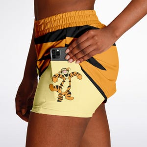 Puede incluir: Pantalones cortos deportivos amarillos y naranjas con rayas negras y un bolsillo con un dibujo animado de Tigger. Los pantalones cortos tienen una cintura con cordón y un teléfono en el bolsillo.