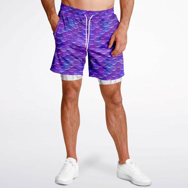 Puede incluir: Pantalones cortos deportivos de color morado y azul con un patr&oacute;n de escamas de sirena. Los pantalones cortos tienen un cord&oacute;n blanco y un forro interior blanco.