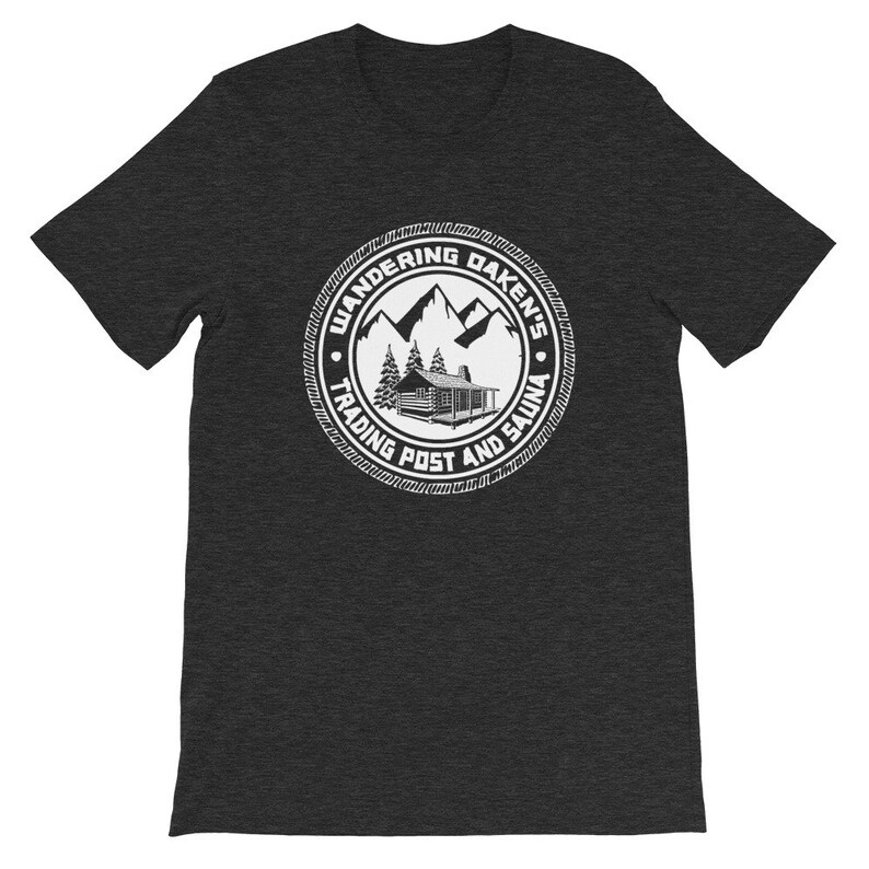 May include: A dark heather grey t-shirt with a white graphic of a cabin in the woods with mountains in the background. The graphic is a circle with the text "Wandering Dakens Trading Post and Sauna".