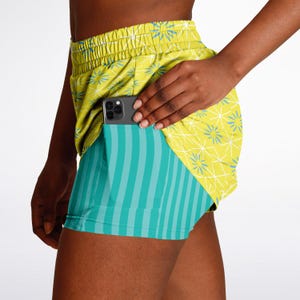 May include: Athletic shorts in yellow and teal, featuring a geometric design and vertical stripes. The shorts have a pocket holding a smartphone. The waistband is elasticated. Designed for running or other sports, they are made for comfort.