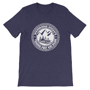 May include: A navy blue t-shirt with a white circular design featuring a cabin, mountains, trees, and the text "Wandering Daken's Trading Post and Sauna".