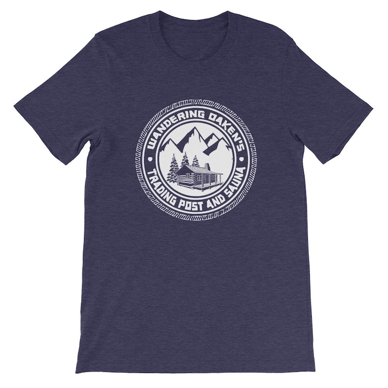 May include: A navy blue t-shirt with a white circular design featuring a cabin, mountains, trees, and the text "Wandering Daken's Trading Post and Sauna".
