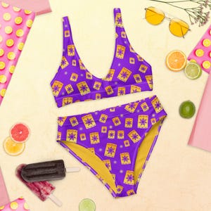 May include: A two-piece swimsuit with a purple background and a repeating pattern of yellow lanterns. The top is a scoop-neck style, and the bottoms are high-waisted. The swimsuit is displayed with sunglasses and fruit.