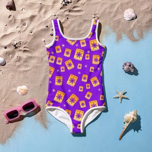 May include: A purple one-piece swimsuit with a pattern of yellow lanterns with sun designs. The swimsuit has white trim and is set on a beach scene with sunglasses, seashells, and a starfish.