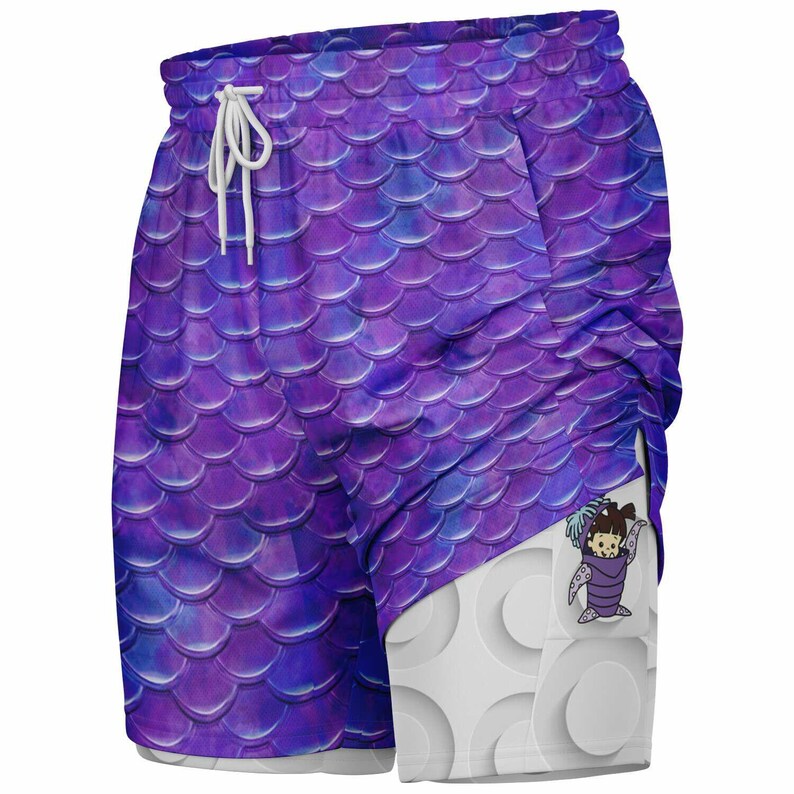 Puede incluir: Pantalones cortos deportivos de color morado con un patr&oacute;n de escamas de sirena y un bolsillo blanco con un personaje de dibujos animados. Los pantalones cortos tienen un cord&oacute;n blanco.