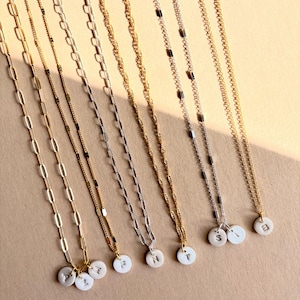 May include: A collection of delicate necklaces with various chain styles, including gold and silver tones. Each necklace features a small, white circular pendant with a single initial. The pendants are arranged in a row, showcasing the personalized jewelry.