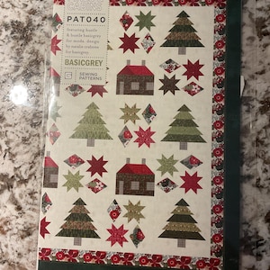 May include: A sewing pattern booklet titled Pines & Cabins PAT040 by BasicGrey. The cover features a quilt design with alternating evergreen trees, small houses, and star shapes in red, green, and cream colors, framed by a floral patterned border.
