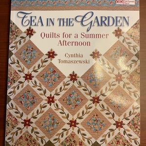 Puede incluir: Un libro de tapa blanda titulado Tea in the Garden: Quilts for a Summer Afternoon de Cynthia Tomaszewski. La portada presenta un patrón de edredón con formas de diamante, cuadrados de tela floral y detalles de flores rojas sobre fondo blanco.