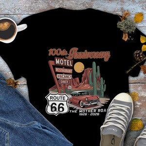 Puede incluir: Camiseta negra con un dise&ntilde;o de la Ruta 66. El gr&aacute;fico incluye un coche antiguo, un paisaje des&eacute;rtico y un letrero de motel, restaurante, habitaciones libres y restaurante. El texto dice "100th Anniversary" y "The Mother Road 1926-2026".