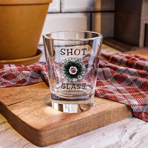 May include: Clear shot glass with the words "SHOT GLASS" in black lettering. A black and red graphic is centered between the words. The shot glass is on a wooden cutting board with a red and white checkered cloth.