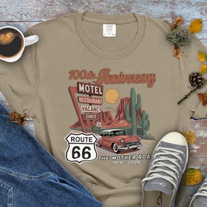 Puede incluir: Camiseta color beige con un dise&ntilde;o de la Ruta 66. El dise&ntilde;o incluye un coche antiguo, un paisaje des&eacute;rtico y un letrero de motel con la inscripci&oacute;n "100th Anniversary". Tambi&eacute;n se ve el texto "The Mother Road 1926-2026".