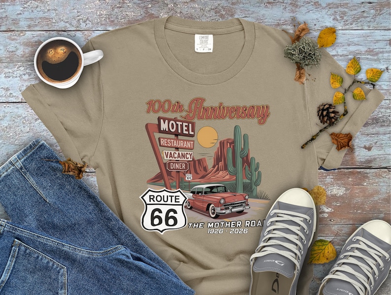 Puede incluir: Camiseta color beige con un dise&ntilde;o de la Ruta 66. El dise&ntilde;o incluye un coche antiguo, un paisaje des&eacute;rtico y un letrero de motel con la inscripci&oacute;n "100th Anniversary". Tambi&eacute;n se ve el texto "The Mother Road 1926-2026".