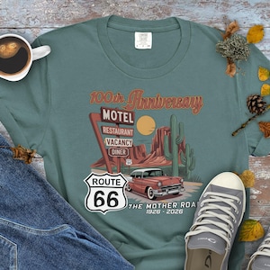 Puede incluir: Una camiseta de color verde azulado con un dise&ntilde;o de la Ruta 66 que incluye un coche antiguo, un letrero de motel y un paisaje des&eacute;rtico. La camiseta conmemora el 100 aniversario de la Ruta 66, con el texto "The Mother Road 1926-2026".