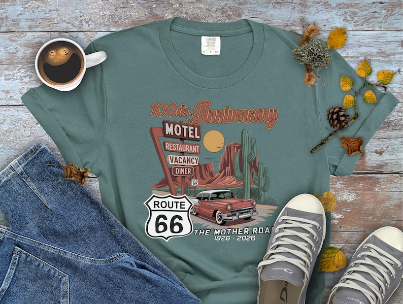 Puede incluir: Una camiseta de color verde azulado con un dise&ntilde;o de la Ruta 66 que incluye un coche antiguo, un letrero de motel y un paisaje des&eacute;rtico. La camiseta conmemora el 100 aniversario de la Ruta 66, con el texto "The Mother Road 1926-2026".