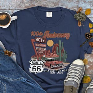 Puede incluir: Camiseta azul marino con un dise&ntilde;o de la Ruta 66. El gr&aacute;fico incluye un coche antiguo, un paisaje des&eacute;rtico y el texto "100th Anniversary Motel". La camiseta tambi&eacute;n tiene el texto "The Mother Road 1926-2026".