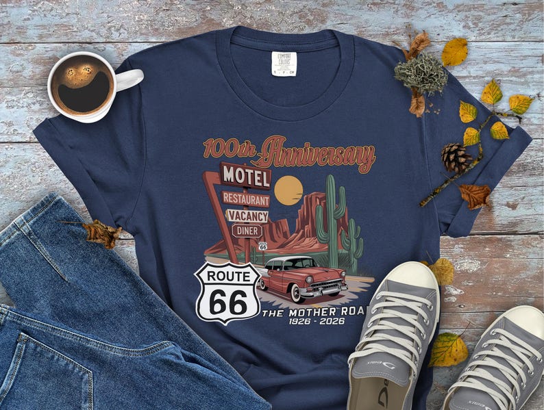Puede incluir: Camiseta azul marino con un dise&ntilde;o de la Ruta 66. El gr&aacute;fico incluye un coche antiguo, un paisaje des&eacute;rtico y el texto "100th Anniversary Motel". La camiseta tambi&eacute;n tiene el texto "The Mother Road 1926-2026".