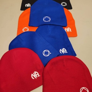 Puede incluir: Una pila de seis gorros de punto en varios colores, incluyendo rojo, naranja y azul. Cada gorro tiene un círculo blanco con un cuadrado en el interior y las letras "NA" en el lateral.
