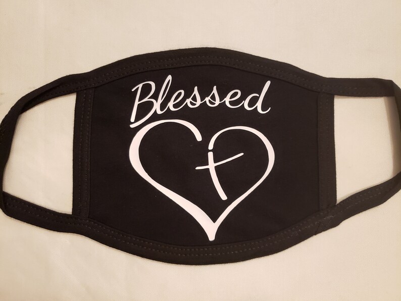 May include: Black face mask with the word "Blessed" in a script font above a white heart with a cross inside. The mask is made of a soft material and has black trim.