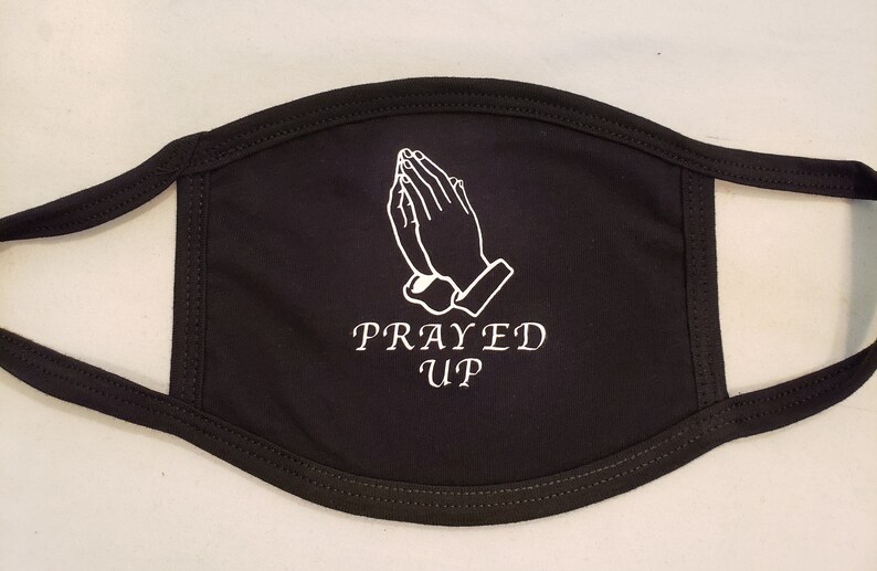 May include: Black face mask with white stitched outline of praying hands and the words "PRAYED UP" in white. The mask has black ear loops and a black border.