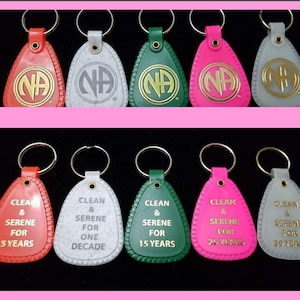 Pode incluir: Dez chaveiros com cores e designs diferentes. Os chaveiros têm a forma de uma lágrima e um anel na parte superior. Os chaveiros têm o texto "CLEAN & SERENE" e o número de anos de sobriedade. As cores dos chaveiros são vermelho, cinza, verde e rosa.