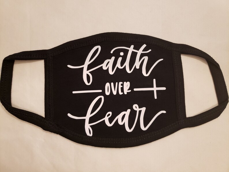 May include: Black face mask with white script that reads "faith OVER fear". The mask has a curved shape to fit the face, with ear loops on each side. The text is centered on the mask.
