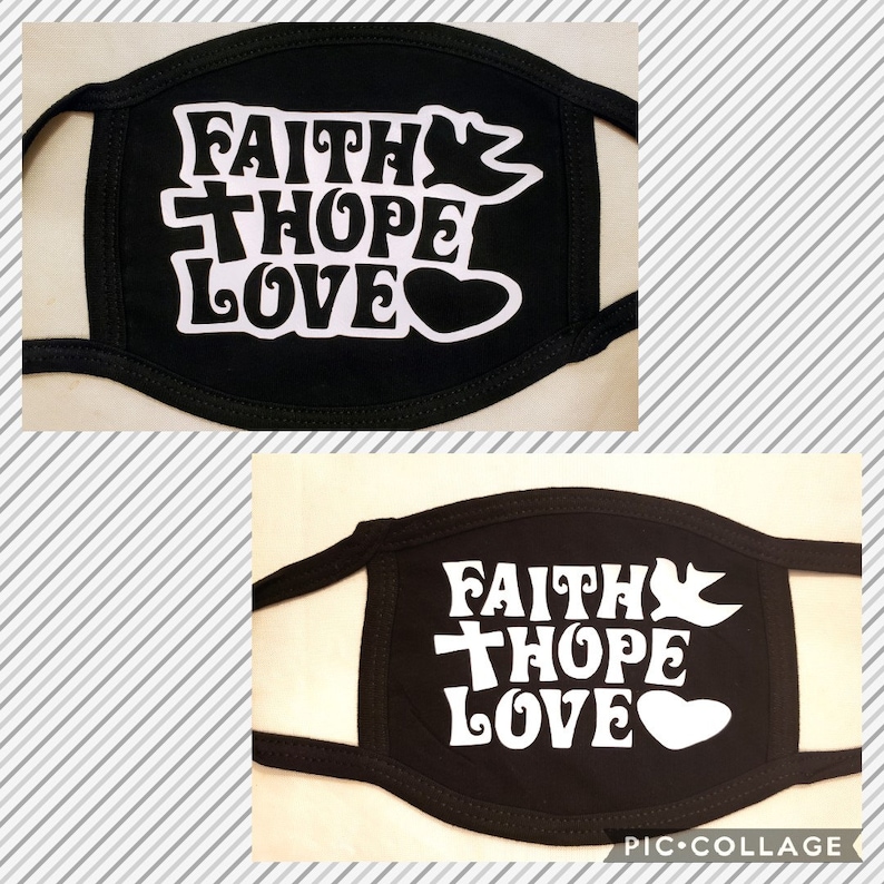 May include: Black face mask with white text that reads "FAITH HOPE LOVE". The design includes a cross, a dove, and a heart. The mask has black ear loops and is set against a striped background.
