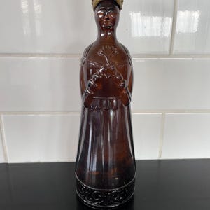 Op de afbeelding: Een bruine glazen fles in de vorm van een persoon die een gouden kroon draagt. De fles heeft ingewikkelde details en een getextureerd oppervlak.