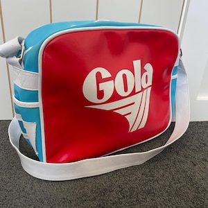以下が含まれることがあります： 鮮やかな赤色のフロントパネルに白いGolaのロゴとグラフィックが施された長方形のショルダーバッグ。側面と上部はライトブルーで、白いパイピングとバッグの側面に取り付けられた白い調節可能なショルダーストラップがアクセントになっています。