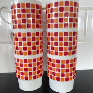 Könnte beinhalten: Vier weiße Keramikbecher, die aufeinander gestapelt sind. Jeder Becher hat ein sich wiederholendes Muster aus roten und orangen Quadraten. Die Becher haben einen Henkel und einen weißen Sockel. Die Becher sind für Heißgetränke konzipiert.