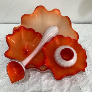 May include: A collection of hand-blown glass art pieces in shades of orange and white. The set includes a large, scalloped-edge bowl, two smaller bowls, and a ladle. The pieces have a glossy finish and are arranged on a white surface.