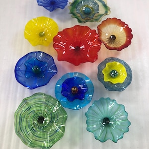 May include: A selection of colourful glass wall art pieces in floral and shell-like designs. The pieces feature vibrant colours including blue, yellow, red, green, and turquoise. Each piece has a unique shape and texture, creating a visually appealing display.