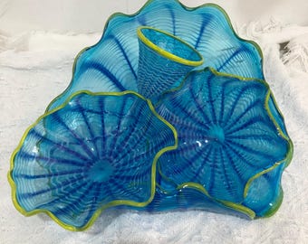 May include: A blue glass sculpture with a wavy, organic shape. The sculpture has a textured surface with a pattern of lines and swirls. The sculpture is accented with a yellow rim.