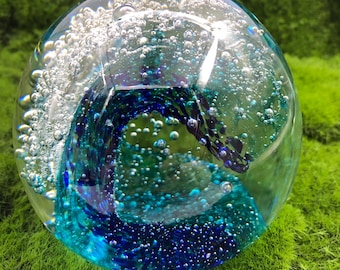May include: A clear glass sphere with a swirling blue and teal design inside. The sphere is filled with many small bubbles, giving it a textured appearance. It sits on a bed of green moss.