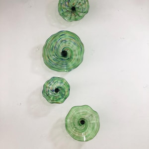 May include: Four green glass wall sculptures with a swirling pattern and a black centre. The sculptures are arranged vertically on a white wall.