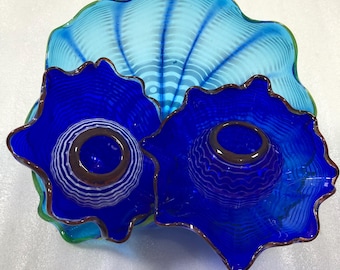 May include: A collection of three decorative glass art pieces. Two are deep blue, bowl-shaped with a brown rim and a central design. The third is a light blue, shell-shaped piece with a green rim and a striped pattern.