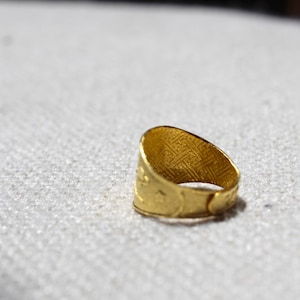 Puede incluir: Un anillo dorado con un dise&ntilde;o intrincado y texturizado. La superficie del anillo presenta un patr&oacute;n repetitivo, y la banda tiene una apariencia detallada y en relieve. El anillo se encuentra sobre un fondo neutro, resaltando su brillo met&aacute;lico.