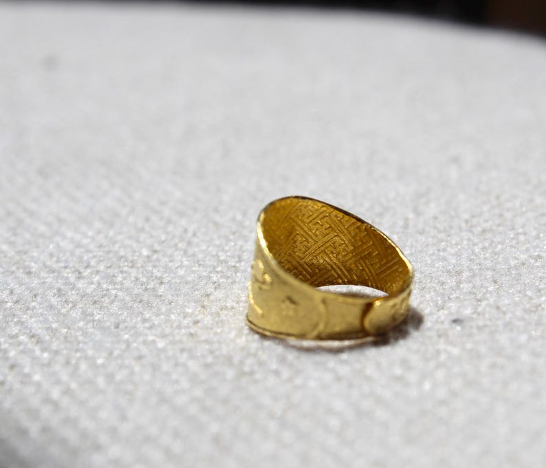 Puede incluir: Un anillo dorado con un dise&ntilde;o intrincado y texturizado. La superficie del anillo presenta un patr&oacute;n repetitivo, y la banda tiene una apariencia detallada y en relieve. El anillo se encuentra sobre un fondo neutro, resaltando su brillo met&aacute;lico.