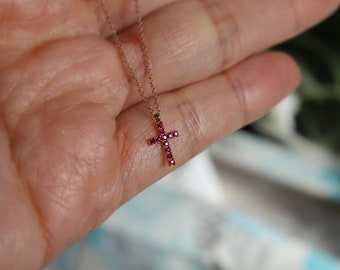 May include: A delicate gold chain necklace with a small cross pendant. The cross is adorned with tiny red gemstones.