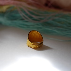 Puede incluir: Un anillo dorado con una cara ovalada y patrones en relieve intrincados. La banda del anillo presenta un dise&ntilde;o de desplazamiento repetido. El fondo incluye hebras de hilo rosa y turquesa, lo que sugiere un tema de artesan&iacute;a o joyer&iacute;a.