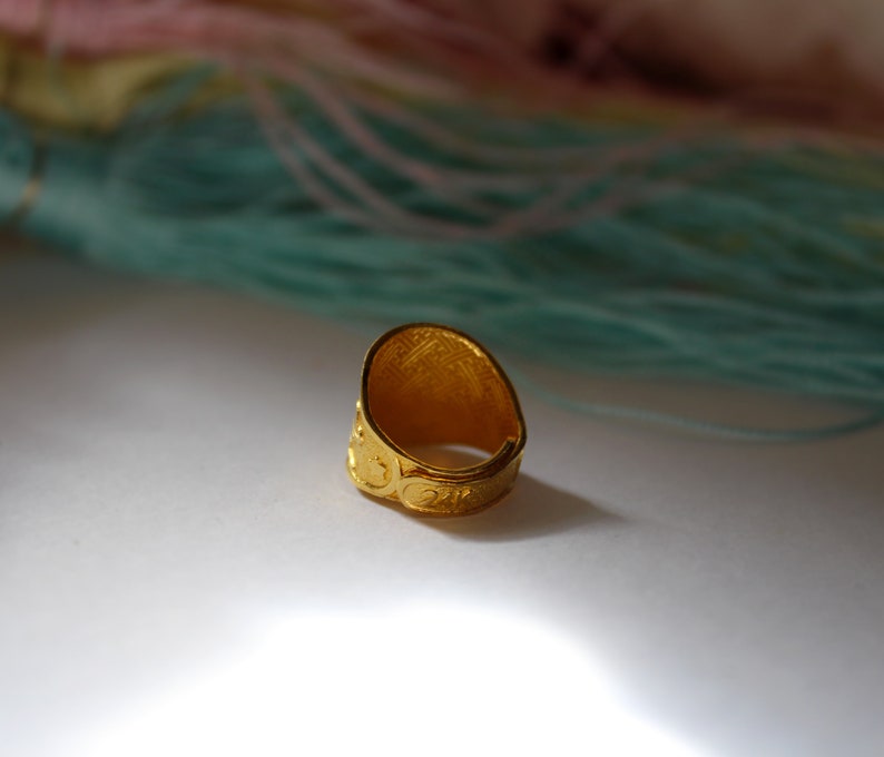 Puede incluir: Un anillo dorado con una cara ovalada y patrones en relieve intrincados. La banda del anillo presenta un dise&ntilde;o de desplazamiento repetido. El fondo incluye hebras de hilo rosa y turquesa, lo que sugiere un tema de artesan&iacute;a o joyer&iacute;a.