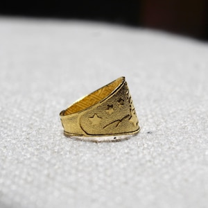 Puede incluir: Un anillo dorado con una superficie texturizada. El anillo presenta una luna creciente, estrellas y un dise&ntilde;o de &aacute;rbol. El anillo es probablemente de metal y se muestra sobre una superficie blanca.