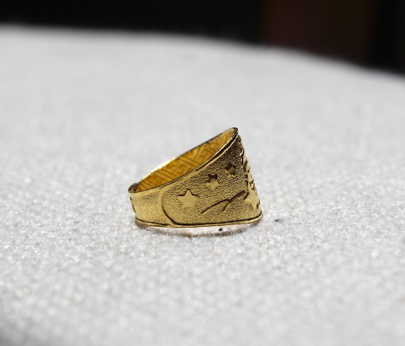 Puede incluir: Un anillo dorado con una superficie texturizada. El anillo presenta una luna creciente, estrellas y un dise&ntilde;o de &aacute;rbol. El anillo es probablemente de metal y se muestra sobre una superficie blanca.