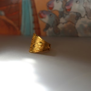 Puede incluir: Un anillo dorado con una superficie texturizada y un dise&ntilde;o que presenta un planeta y otros elementos celestes. El anillo tiene una banda ancha y una forma ligeramente triangular, con intrincados patrones calados.