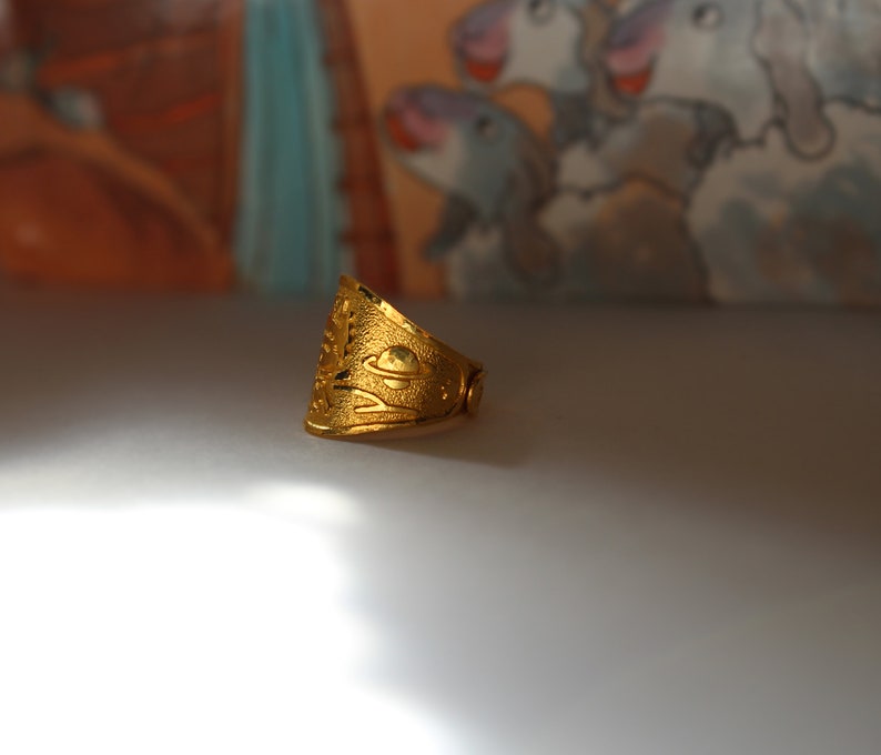 Puede incluir: Un anillo dorado con una superficie texturizada y un dise&ntilde;o que presenta un planeta y otros elementos celestes. El anillo tiene una banda ancha y una forma ligeramente triangular, con intrincados patrones calados.