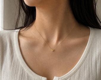 Puede incluir: Un delicado collar de oro con un pequeño colgante en forma de corazón. El collar se lleva puesto y la cadena de oro es visible sobre la piel. El colgante de corazón está centrado en el pecho.