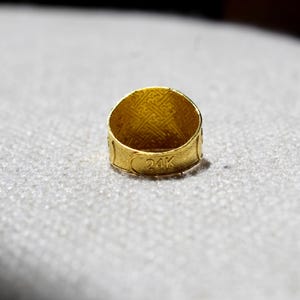 Puede incluir: Primer plano de un anillo de oro con el texto "24K" grabado en la banda. El anillo tiene un interior texturizado y un patr&oacute;n decorativo en el exterior. El anillo est&aacute; sobre un fondo claro y texturizado.