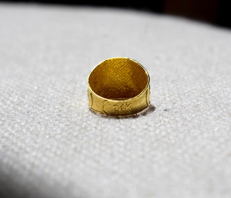 Puede incluir: Primer plano de un anillo de oro con el texto "24K" grabado en la banda. El anillo tiene un interior texturizado y un patr&oacute;n decorativo en el exterior. El anillo est&aacute; sobre un fondo claro y texturizado.