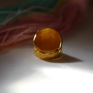 Puede incluir: Primer plano de un anillo de oro con superficie texturizada. El anillo est&aacute; inscrito con "14K" y presenta un dise&ntilde;o detallado alrededor de la banda. El anillo se encuentra sobre un fondo suave y borroso con tonos rosas y verdes.